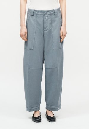 Pantalon gris à jambe droite avec poches plaquées avant, détails de couture aux genoux et poignets boutonnés, porté avec des ballerines noires et un haut blanc.