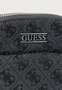 Μαύρο πορτοφόλι με υφή, διακοσμημένο με μοτίβο μονόγραμμα, ασημένιο λογότυπο "GUESS" και ορατό μεταλλικό φερμουάρ.