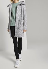 Osoba oblečená v svetle šedém plášti s kapucí, mátově zeleném svetru, černých kalhotách a bílých teniskách, kráčející proti jednoduchému šedému pozadí.