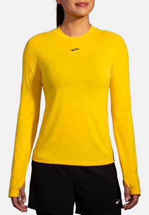 Kvinde iført en knaldgul atletiktrøje med lange ærmer og tommelfingerhuller samt sorte atletikshorts mod en ensfarvet baggrund.