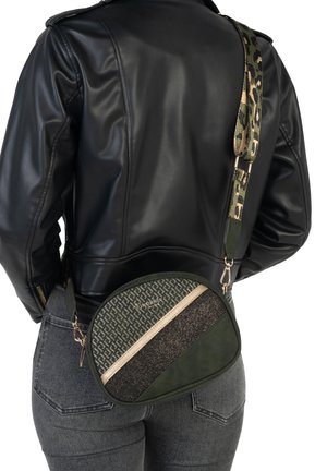 Une personne portant une veste en cuir noir et un jean gris porte un sac bandoulière oval en vert à motifs avec une sangle camouflage.