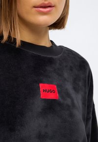 Sweatshirt de veludo preto com colarinho canelado, apresentando um patch retangular bordado em vermelho com "HUGO" em texto preto.