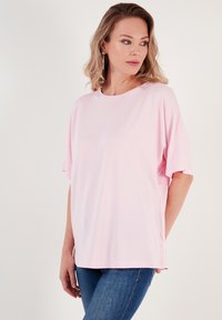 Světle růžové oversized tričko s krátkými rukávy, kulatým výstřihem, měkkou tkaninou a postranními rozparky pro snadný pohyb. Na sobě má modré džíny.