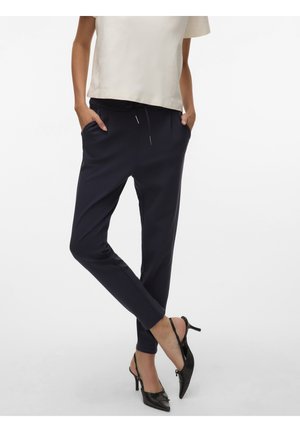 Vrouw die marineblauwe taps toelopende broek met zijzakken draagt, een crèmekleurige top met korte mouwen, en zwarte slingbacks met spitse neus, staand met gekruiste benen.