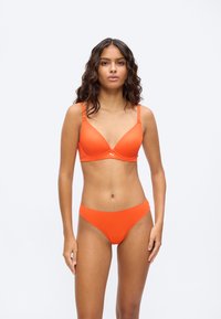 Γυναίκα με κυματιστά σκούρα μαλλιά, φοράει φωτεινό πορτοκαλί σουτιέν και σλιπ που ταιριάζουν, στέκεται μπροστά σε απλό φωτεινό φόντο.