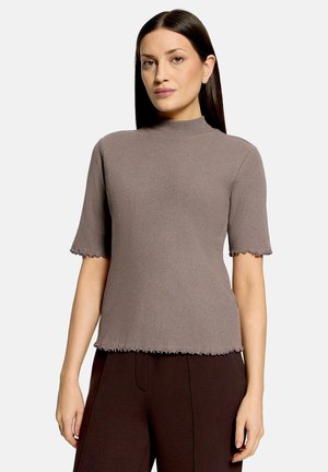 Woman with straight dark hair wears a taupe short-sleeve ribbed top with frilled edges and dark brown pants, standing against a plain background.