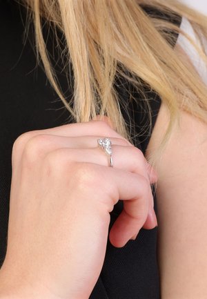 Zilveren ring met een peervormige heldere steen, met een dunne band. De hand is gepositioneerd tegen een zwarte achtergrond, met zichtbare lichtreflecties.