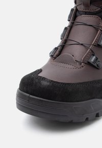 Pruun ja must matkajalatseid tugevdatud varbaga, karm tallaga ja kiire nöörimise süsteemiga, näidatud lihtsa valge tausta ees.