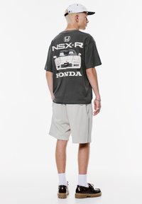 Ung mand iført en sort Honda NSX-R grafisk T-shirt, lysegrå shorts, hvide sokker, brune sko og en hvid baseballcap, som står sidelæns.