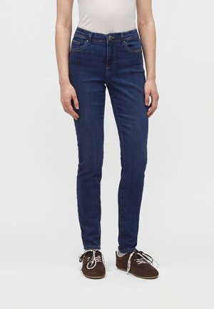 Personne portant un jean slim bleu foncé, un haut blanc et des chaussures marron à lacets, debout devant un fond blanc uni.