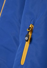 Blaues Stoffjackett mit einer gelben Reißverschlusstasche und einem schwarzen Reißverschlussanhänger. Die glatte Textur und die klaren Designdetails sind sichtbar.