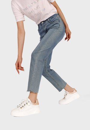 Persona con jeans azzurri chiari e sneakers platform bianche con dettagli beige, posando davanti a uno sfondo chiaro e uniforme.