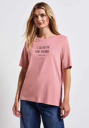 Lyserød t-shirt med korte ærmer og rund halsudskæring. Har sort tekst: "RO PÅ INSIDEN, FOKUSER PÅ DE STILLE SANSE."