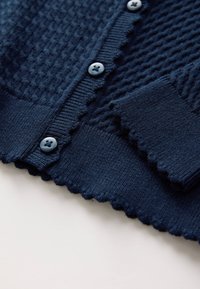 Marineblauwe gebreide cardigan met een gestructureerd patroon, geschulpte randen en blauwe knopen. Zachte stof met geribbelde manchetten en zoom.