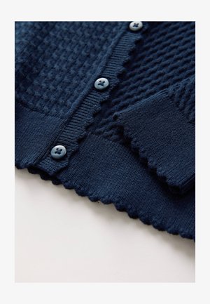 Tumši zils adīts kardigans ar fakturētu rakstu, viļņotām malām un zilām pogām. Mīksts audums ar ribotiem aprocēm un apakšu.