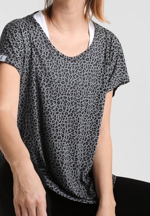 Vrouw draagt een grijze top met korte mouwen met een zwart luipaardprint-patroon, gelaagd over een wit hemdje, zittend tegen een effen achtergrond.