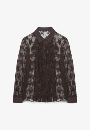 Blusa in pizzo marrone con colletto marrone scuro, caratterizzata da dettagli arricciati sulla parte anteriore e chiusura con bottoni. Tessuto trasparente con motivi floreali.