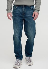 Jeans de culoare albastru închis, cu un model drept, cinci buzunare și ușoare deteriorări. Asortați cu pantofi sport gri. Purtați cu un pulover din tricot.
