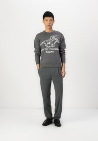 Grijze trui met een witte grafische print en de tekst "MUIS RENDE WEG." Gecombineerd met grijze broek met streepjes en zwarte schoenen.