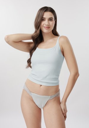 Mladá žena v světle modrém tílku a odpovídajícím spodním prádle s krajkovým lemem, usmívající se s jednou rukou na krku, na jednobarevném pozadí.