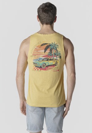 Hombre vistiendo una camiseta sin mangas amarilla con un gráfico de coche vintage, palmeras y puesta de sol, combinado con pantalones cortos de mezclilla azul claro.