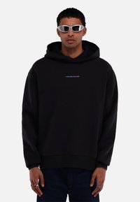 Zwarte katoen hoodie met een voorste kangoeroezak, met een "TOMORROWLAND" logo in blauw en roze. Model draagt reflecterende zonnebril.