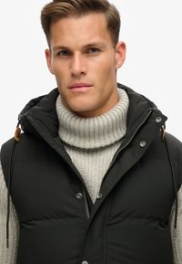 Schwarze Weste aus Puffmaterial mit Kapuze, ausgestattet mit silbernen Druckknöpfen und Kordeln, über einem hellgrauen gestreiften Rollkragenpullover getragen.