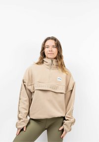 Beige Fleece-Pullover mit Viertelreißverschluss und hohem Kragen, der ein strukturiertes Logo und gestickten Text aufweist, lange Ärmel mit Akzenten, getragen mit olivfarbenen Leggings.
