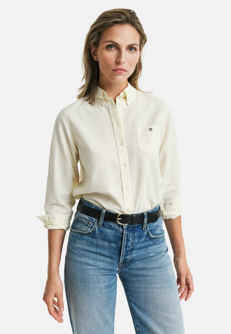 Light yellow button-up shirt with a chest pocket, rolled sleeves, paired with blue denim jeans and a black belt with a round buckle.