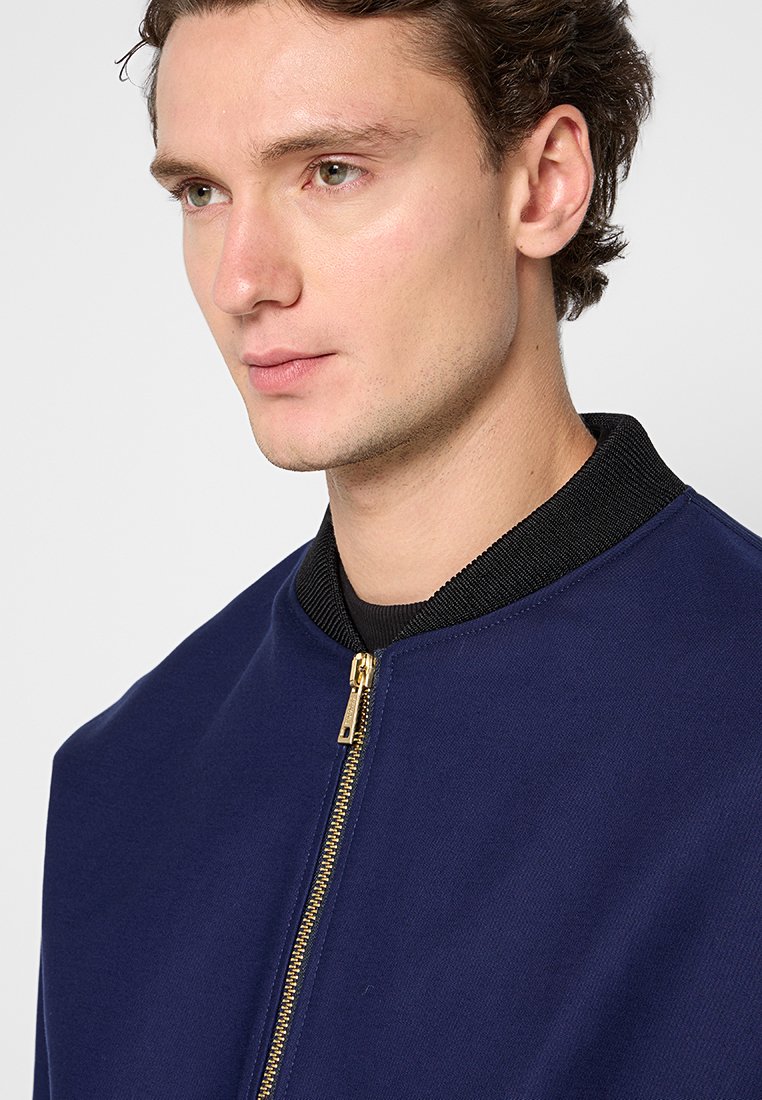 Marine Bomberjacke aus glattem Stoff, mit einem schwarzen Rippkragen und einem goldenen Reißverschluss, in einer schlichten, strukturierten Silhouette.