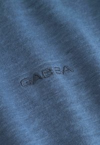 Blått tyg med en slät textur som har en präglad "GABBA"-logotyp i en kontrasterande färg. Fokus på stickstrukturen.