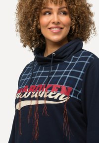 Felpa navy con un motivo a scacchi, grafica prominente "UNBROKEN" in rosso e dettagli decorativi arancioni lungo l'orlo. Materiale in morbido misto cotone.