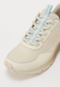 Zapatilla deportiva beige con una parte superior texturizada, cordones azul claro y una suela con un diseño estampado. Cuenta con un diseño acolchado y una cinta en el talón.