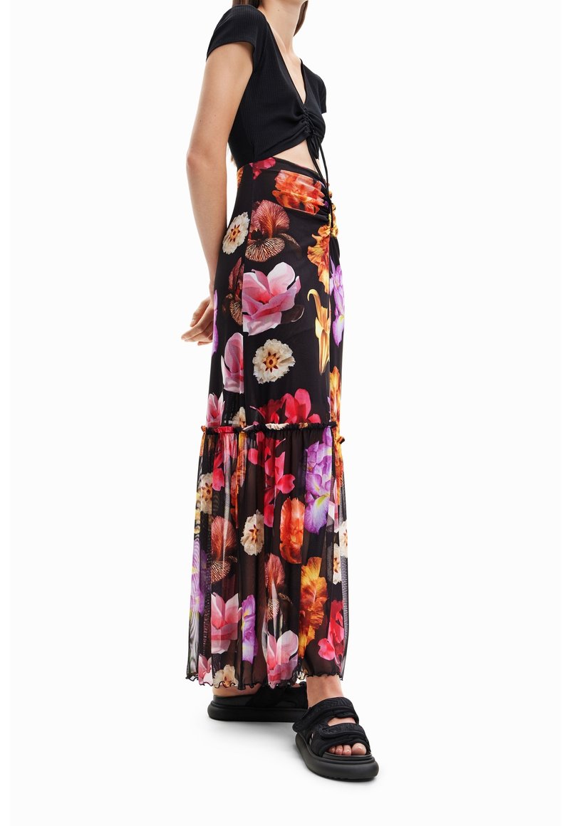Jupe maxi avec un motif floral vibrant sur un tissu noir, dotée d'un ourlet volanté et d'une taille élastique. Assortie à un top noir noué et des sandales.
