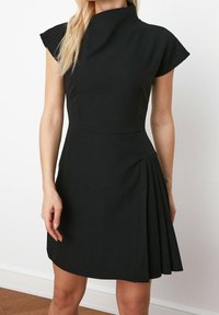 Une femme porte une robe noire avec un col haut et des manches courtes, présentant un design plissé sur le côté, se tenant dans un espace intérieur bien éclairé.