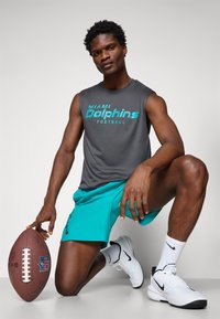 Graues ärmelloses Shirt mit "Miami Dolphins Football" in Türkis, kombiniert mit türkisen Shorts. Weiße Sneakers und ein brauner Football sind vorhanden.