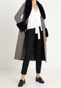 Vrouw in een geruite lange jas met zwarte bontkraag en -manchetten, witte wikkelblouse, zwarte slim-fit broek en zwarte instappers, staande op een witte vloer.