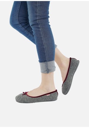 Ben i opkørte blå jeans iført grå filt-slip-on-tøfler med burgunderrøde kanter og små sløjfer på en hvid baggrund.