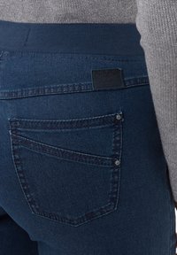 Jeans blu scuro con una texture liscia, dotati di una tasca posteriore curva, un'etichetta del marchio e una vita a coste. Dettagli di cucitura sottili sono visibili.