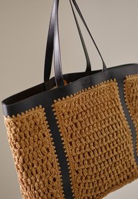 Velika torba za na rame s ravnimi ročaji je opredeljena z rjavkasto kvačkanimi predeli in črnimi usnjenimi poudarki, ima teksturirano površino in odprt dizajn.