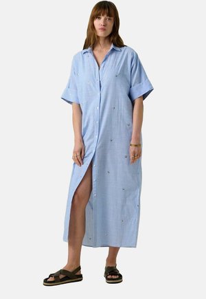 Robe chemise bleu et blanc à rayures avec des manches courtes, présentant des accents décoratifs et une fente latérale, associée à des sandales sombres.