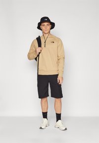 Hnedá mikina s kapucňou, čierny vedierkový klobúk, čierne krátke nohavice a biele tenisky. Batoh držaný v pravej ruke. Jednoduchý, neformálny outfit.