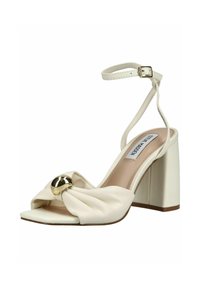 Sandalia crema de tacón bloque abierto con correa en el tobillo, correa frontal torcida y gran adorno decorativo dorado en la pala.