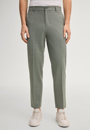 Mężczyzna ubrany w oliwkowe, dopasowane spodnie wizytowe i białe trampki, stojący na jednolitym tle.