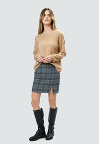 Beige Strickpullover mit strukturiertem Einsatz, kombiniert mit einem grauen karierten Rock mit einem Schlitz auf der Vorderseite. Schwarze kniehohe Stiefel runden den Look ab.