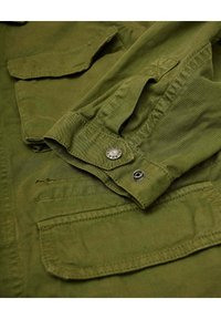 Chaqueta verde oliva de tela texturizada, con puños abotonados y múltiples bolsillos en la parte frontal para almacenamiento.