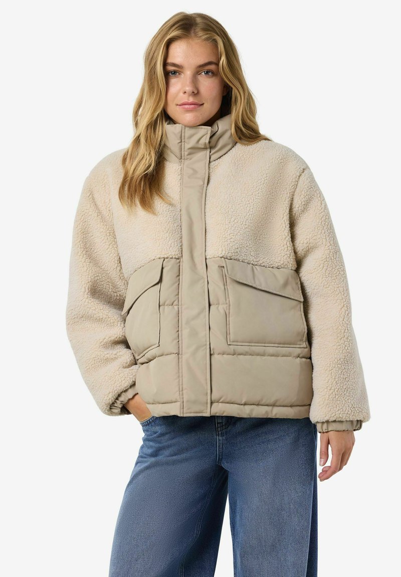 Kobieta w beżowej kurtce z wysokim kołnierzem z sherpy i pikowanej, z dużymi kieszeniami z przodu i niebieskimi dżinsami, stojąca z jedną ręką w kieszeni.