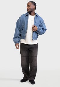 Farmert bomber dzseki laza szabással, világoskék színben, bordázott mandzsettákkal és rugalmas szegéllyel, sötét, oversized farmernadrággal és fekete cipőkkel párosítva.