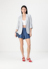 Juostuotas švarkas baltos ir mėlynos spalvos, baltas crop top'as, mėlyni taškeliai šortai ir raudoni aukštakulniai su metaliniais akcentais, pozavimas prieš paprastą foną.