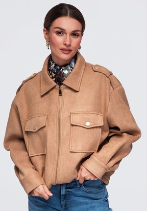 Femme portant une veste beige zippée avec de grandes poches devant et une écharpe à motifs sous le col, posant avec les mains près de la taille.